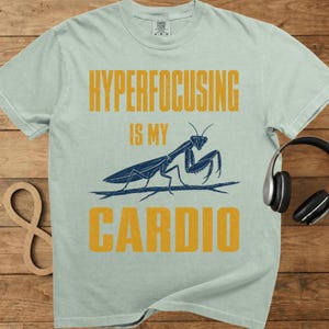 May include: Light blue t-shirt with the text "HYPERFOCUSING IS MY CARDIO" in yellow. A blue illustration of a praying mantis is below the text. The shirt has a crew neck and short sleeves.