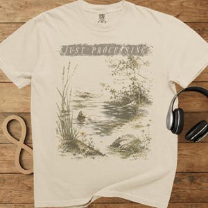May include: A cream-colored short-sleeve t-shirt featuring a graphic print of a calm lake scene with tall grasses, a small tree, and a rock. The text "JUST PROCESSING" is printed in a distressed font above the illustration on the front.