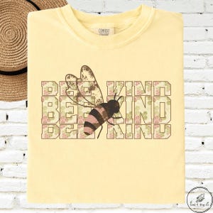 May include: A light yellow t-shirt with the words "BEE KIND" in a block letter design. A bee graphic is centered on the shirt, with brown and pink stripes. The shirt is folded on a white brick wall.