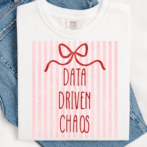 Puede incluir: Camiseta blanca con un lazo rojo y las palabras "DATA DRIVEN CHAOS" en rojo. El fondo presenta rayas verticales rosas y blancas. La camiseta está doblada sobre mezclilla azul.