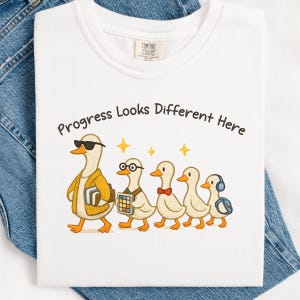 Peut inclure: T-shirt blanc avec l'inscription « Progress Looks Different Here » et un dessin de cinq canards. Le premier canard porte des lunettes de soleil et une veste jaune, le second porte des lunettes et un Rubik's Cube.