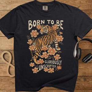 Op de afbeelding: Zwart T-shirt met een tijgerillustratie in oranje en zwart, omringd door oranje bloemen. De tekst "BORN TO BE GLORIOUSLY UNSCRIPTED" staat boven en onder het tijgerontwerp. Het shirt heeft een ronde hals.