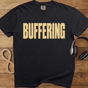 May include: Black t-shirt with the word "BUFFERING" printed in a light tan color across the front. The shirt has a crew neck and short sleeves. A pair of black headphones rests on the right side of the shirt.