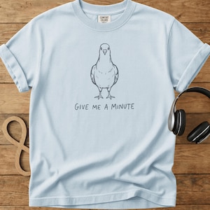 May include: Pale blue sweatshirt featuring a pigeon illustration and the phrase "GIVE ME A MINUTE". The design is in a handwritten style. The sweatshirt has a crew neck and rolled sleeves. The brand "COMFORT COLORS" is printed on the tag.