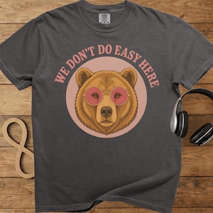 以下が含まれることがあります： ピンクのサングラスをかけた茶色のクマが描かれた、濃いグレーのTシャツ。ピンクの円の中にクマが配置され、周りに「WE DON'T DO EASY HERE」の文字が弧を描いています。Tシャツはクルーネックで半袖です。