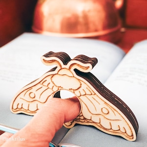 Peut inclure: Une bague en forme de papillon de nuit en bois, avec un motif d'ailes détaillé, posée sur un livre ouvert. La bague est en bois brun clair et foncé. L'arrière-plan comprend une bouilloire en cuivre et des meubles en bois.