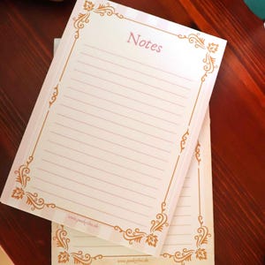 Könnte beinhalten: Ein rechteckiger Notizblock aus weißem Papier mit hellrosa horizontalen Linien und einem goldfarbenen Zierrand. Das Wort Notes ist oben mittig in Rosa gedruckt, und die Website www.punktilue.de ist unten aufgedruckt.