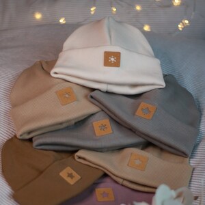 Può includere: Una pila di sei berretti di lana di diversi colori, tra cui beige, marrone, grigio e rosa. Ogni berretto ha una piccola toppa quadrata con un design diverso, tra cui un fiocco di neve, una stella e una foglia d'acero.