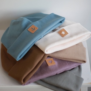 May include: A stack of five knitted beanies in different colours, including blue, white, brown, purple, and grey. Each beanie has a small leather patch with a different design, including an anchor, a star, and a heart.