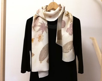 May include: A white silk scarf with a floral print of leaves and flowers in shades of brown and beige. The scarf is hanging on a wooden hanger against a white wall.