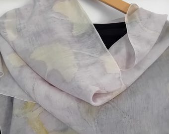Può includere: Una sciarpa trasparente grigio chiaro con un motivo simile a un acquerello di pennellate giallo pallido e bianco. La sciarpa è drappeggiata su un appendiabiti di legno, rivelando un capo nero sottostante. Il tessuto appare morbido e fluido.