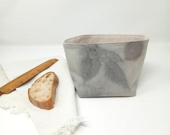 May include: A square-shaped, light gray fabric storage basket with a natural leaf print design. The basket is made of linen and has a light pink lining. The basket is sitting on a white surface.