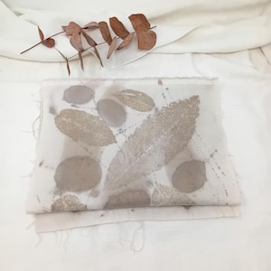 May include: A folded white fabric with leaf imprints in shades of brown and gray. The fabric has a raw edge and is decorated with various leaf shapes and patterns. A sprig of dried eucalyptus leaves rests above the fabric.
