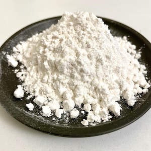 Può includere: Un cumulo di polvere bianca, possibilmente un ingrediente alimentare o un prodotto cosmetico, è posto su un piatto verde scuro. La polvere appare finemente macinata con una superficie leggermente strutturata. Il bordo del piatto è visibile.