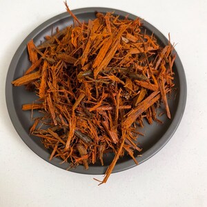 Può includere: Un piatto grigio scuro pieno di corteccia sminuzzata arancione-marrone. I pezzi di corteccia sono di varie lunghezze e spessori, creando un aspetto naturale e strutturato. Il piatto è rotondo e poggia su una superficie bianca.