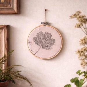 Pode incluir: Um bastidor de bordado redondo de madeira com um tecido rosa claro com uma grande impressão de folha cinza. O bastidor é suspenso por uma corda marrom. Decoração de parede com tema natural.