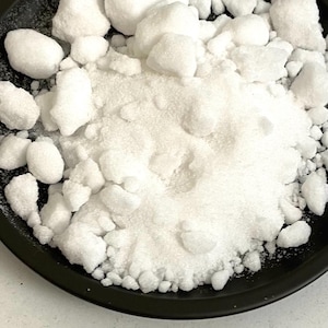 Può includere: Primo piano di sostanze cristalline bianche, tra cui pezzi grossolani e granuli fini, su un piatto nero. Le trame variano da forme grandi e irregolari a una consistenza polverosa. L'immagine suggerisce un ingrediente alimentare o un composto chimico.