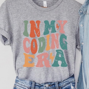 May include: A gray t-shirt with the text "In My Coding Era" in colorful retro-style letters. The letters are outlined in white and have a distressed look.