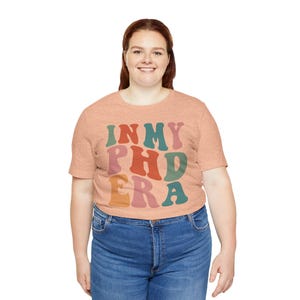 以下が含まれることがあります： 桃色のTシャツに「IN MY PHD ERA」というレトロなフォントでカラフルな文字が書かれています。文字は緑、オレンジ、赤、ピンクの色合いです。
