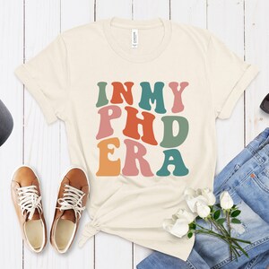 以下が含まれることがあります： ベージュのTシャツに、レトロなスタイルのカラフルな文字で「In My PhD Era」と書かれています。