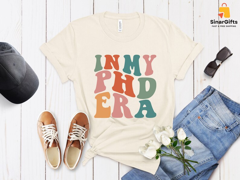 以下が含まれることがあります： ベージュのTシャツに、レトロなスタイルのカラフルな文字で「In My PhD Era」と書かれています。