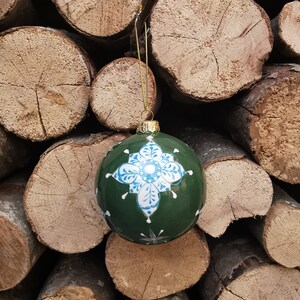 May include: A green Christmas bauble with a white and blue snowflake design. The bauble is hanging from a gold string and is resting on a pile of stacked logs.