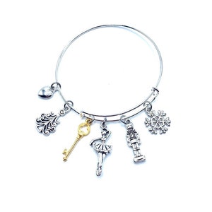 May include: Silver-toned bangle bracelet with multiple charms. Charms include a heart, a decorative flourish, a gold-colored key, a ballerina, a nutcracker, and a snowflake. The bracelet is on a white background.