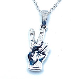 May include: Silver-toned pendant necklace featuring a hand gesture forming a peace sign. The pendant is detailed with visible fingers and attached to a delicate chain. The necklace is displayed against a white background.