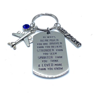 May include: Silver-toned keychain with a dog tag pendant, airplane charm, and "AIR FORCE" charm. The dog tag has the text "ALWAYS REMEMBER YOU ARE BRAVER THAN YOU BELIEVE..." A blue gem is attached to the keychain.