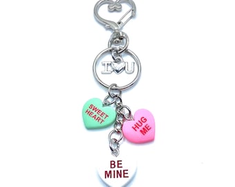 Pode incluir: Um porta-chaves prateado com um fecho em forma de coração e um anel com as letras "I U". Apresenta três pingentes em forma de coração em verde, rosa e branco. Cada pingente tem uma mensagem diferente em texto vermelho: "SWEET HEART", "HUG ME" e "BE MINE".