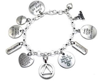 May include: A silver-toned charm bracelet featuring multiple circular, rectangular, and heart-shaped pendants. The charms are engraved with phrases including One day at a time, Never give up, WillPower, WARRIOR, and It's a beautiful day to be sober.