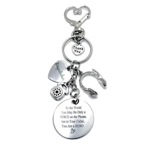 May include: Silver-toned keychain with multiple charms. Charms include a heart with "Thank You", a heart with "Dispatcher", a telephone, and a headset. A circular charm reads "To the World You May Be Only a VOICE on the Phone, but to Your Caller, You Are a HERO".
