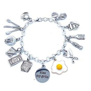 May include: A silver charm bracelet featuring various food-related charms. Charms include a fried egg, a slice of toast, a $100 bill, and a charm that says "kiss my grits". Other charms include a fork, knife, and spoon.