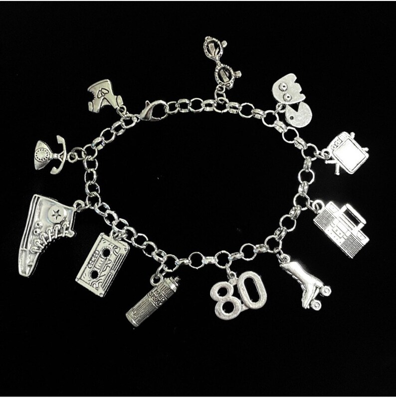 May include: A silver-toned charm bracelet featuring various miniature pendants including a high-top sneaker, a cassette tape, a hairspray can, the number 80, a roller skate, a boombox, a television, a ghost-like figure, glasses, and a telephone.