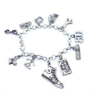 May include: A silver-toned charm bracelet featuring various miniature pendants including a high-top sneaker, a cassette tape, a hairspray can, the number 80, a rotary telephone, roller skates, a boombox, a television, and eyeglasses.