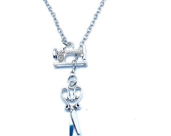 May include: Silver-toned necklace featuring a sewing machine charm and a pair of scissors charm. The sewing machine charm has detailed embellishments. The scissors have blue accents on the blades. The necklace is on a white background.