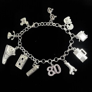 May include: A silver-toned charm bracelet featuring various miniature pendants including a high-top sneaker, a cassette tape, a hairspray can, the number 80, a roller skate, a boombox, a television, a ghost-like figure, glasses, and a telephone.