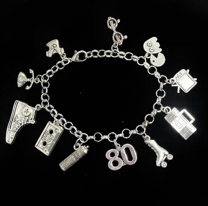 May include: A silver-toned charm bracelet featuring various miniature pendants including a high-top sneaker, a cassette tape, a hairspray can, the number 80, a roller skate, a boombox, a television, a ghost-like figure, glasses, and a telephone.
