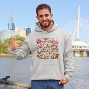 Puede incluir: Sudadera con capucha gris claro con un colorido collage de iconos canadienses. El diseño incluye una hoja de arce, una taza de café, un castor y el texto "eh?". La sudadera con capucha la lleva una persona frente a un fondo escénico.