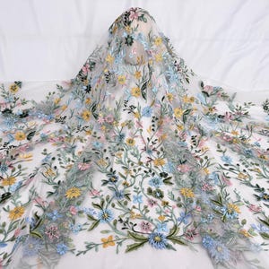 Könnte beinhalten: Weißer Tüllstoff mit farbenfroher Blumenstickerei. Die Stickerei zeigt gelbe, blaue und rosa Blumen mit grünen Blättern und Stielen. Der Stoff ist über einer unsichtbaren Form drapiert.