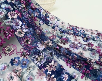 Puede incluir: Primer plano de una tela transparente con diseños florales bordados. La tela presenta una base azul marino con intrincados patrones de flores moradas, rosas y azules, adornadas con lentejuelas. El bordado floral es detallado y en capas.