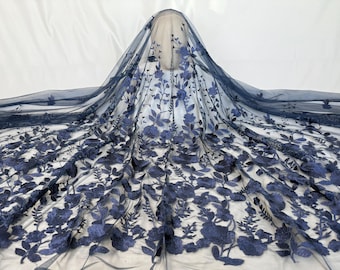 May include: Navy blue floral embroidered tulle fabric. The fabric features a delicate pattern of blue flowers and leaves. The tulle is draped over a white surface, showcasing the intricate design. Ideal for bridal wear, dresses, or decorative projects.