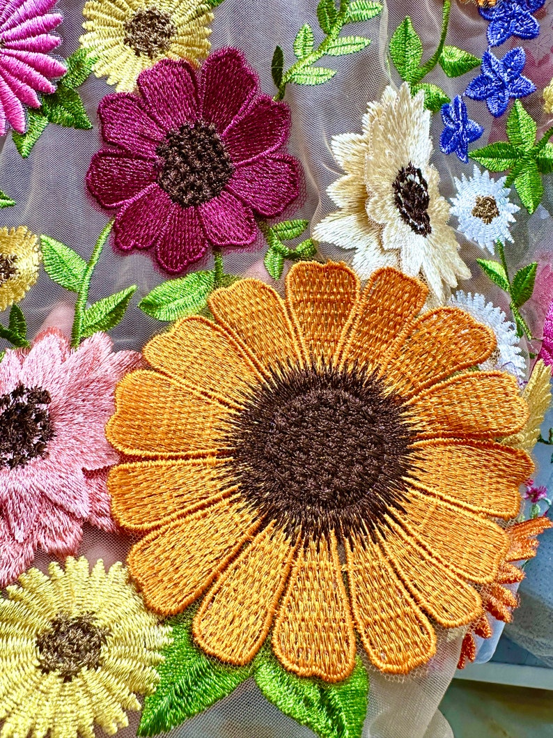 May include: A close-up of a fabric with embroidered flowers. The flowers are in shades of pink, yellow, orange, and blue. The fabric is a sheer mesh.