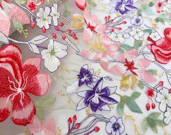 May include: Close-up of embroidered floral fabric. The design features a variety of flowers in shades of red, pink, purple, and white, with green leaves and stems. The embroidery is detailed and delicate, creating a textured effect.