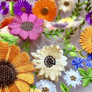 May include: A sheer fabric with embroidered flowers in various colors, including pink, yellow, orange, white, and purple. The flowers are arranged in a random pattern and are surrounded by green leaves.