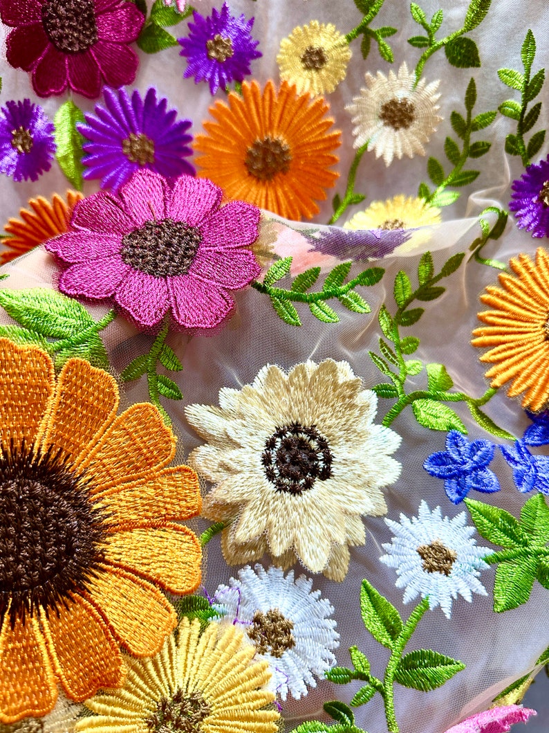 May include: A sheer fabric with embroidered flowers in various colors, including pink, yellow, orange, white, and purple. The flowers are arranged in a random pattern and are surrounded by green leaves.
