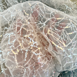 Puede incluir: Tela blanca transparente con un delicado patrón de lentejuelas plateadas. Las lentejuelas están dispersas por toda la tela, creando un efecto brillante.