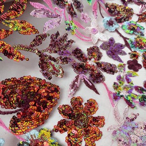 May include: Detailed shot of a fabric with floral embroidery. The design incorporates colourful sequins in red, orange, yellow, green, purple, and pink, forming a textured and lively pattern. The fabric is likely for decorative use, such as embellishments.