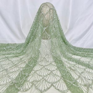 Puede incluir: Una tela verde pálido con un acabado brillante e iridiscente. La tela tiene un delicado patrón de escamas y está adornada con pequeñas cuentas brillantes.