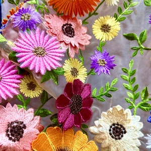 May include: A sheer fabric with embroidered flowers in various colors, including pink, purple, yellow, orange, and red. The flowers are arranged in a random pattern and are surrounded by green leaves.